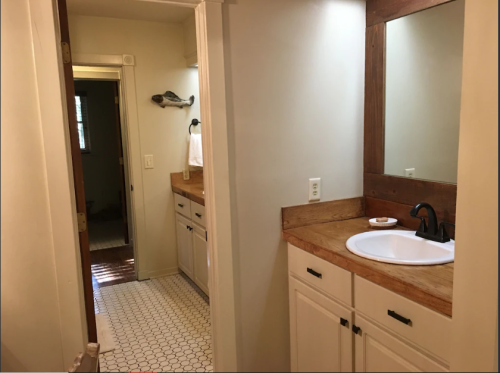 A bright bathroom with a wooden countertop, sink, mirror, and white cabinets, viewed from a hallway.