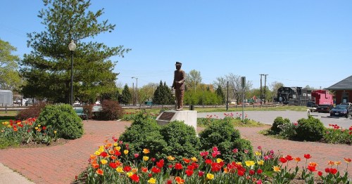 A bronze statue of a soldier stands in a park surrounded by colorful tulips and greenery under a clear blue sky.