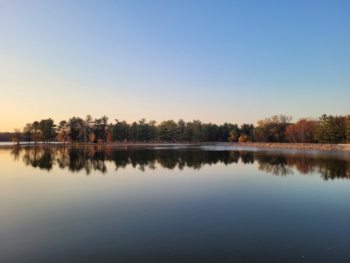 A serene lake at sunset, reflecting trees and a clear sky, with autumn colors in the background.
