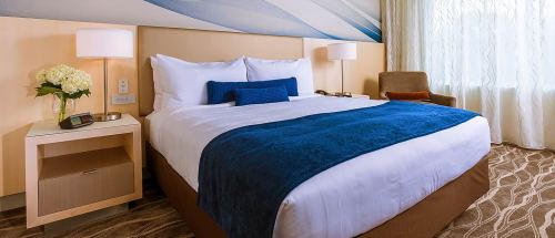 A cozy hotel room featuring a large bed with blue accents, bedside lamps, and a comfortable chair.