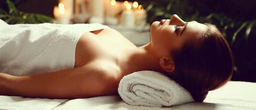 A woman relaxes on a spa table, covered with a towel, surrounded by candles and greenery.