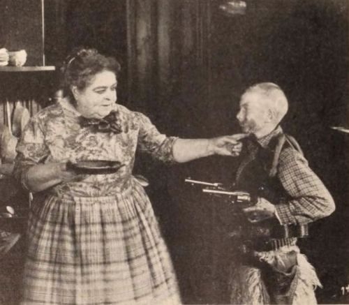 A woman in a dress holds a plate while a small man in cowboy attire points a toy gun at her.