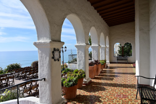 A coastal view from a covered terrace with arches, potted plants, and a patterned floor, overlooking the ocean.