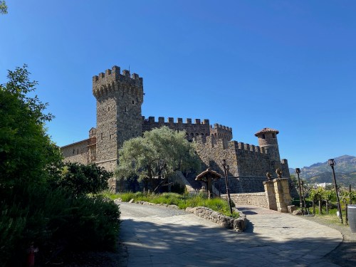 A stone castle with towers and a pathway, surrounded by greenery and mountains under a clear blue sky.
