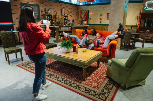 A woman takes a photo of two friends lounging on a couch in a cozy, colorful living room setting.