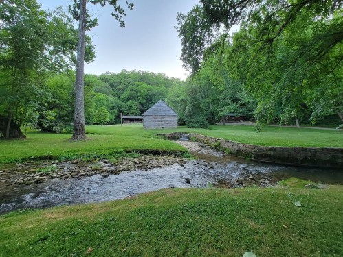 A serene landscape featuring a rustic barn by a stream, surrounded by lush green trees and grass.