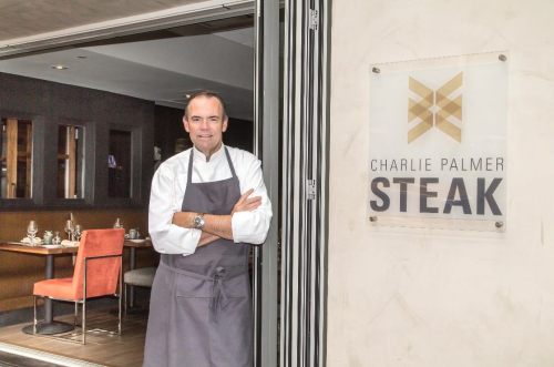 Chef standing at the entrance of Charlie Palmer Steak restaurant, smiling with arms crossed beside the restaurant sign.
