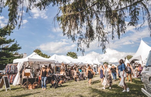 A bustling outdoor market with white tents, shoppers browsing, and a blue sky with scattered clouds overhead.