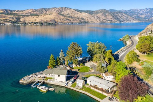 Aerial view of a serene lakeside property with boats, trees, and mountains in the background under a clear blue sky.
