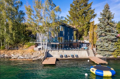 Modern lakeside house with a deck, surrounded by trees and a floating trampoline in the water. Clear blue sky above.
