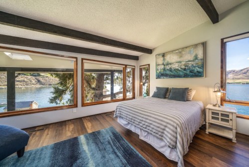 Cozy bedroom with a striped bedspread, large windows, and a view of a lake surrounded by trees.