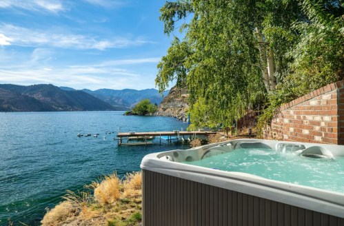 A hot tub by a serene lake, surrounded by mountains and trees under a clear blue sky.