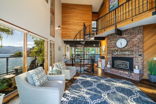 Cozy living area with large windows, a fireplace, and a spiral staircase, overlooking a scenic lake view.