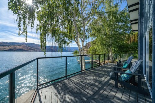A serene lakeside view from a deck with two chairs, surrounded by trees and mountains under a clear blue sky.