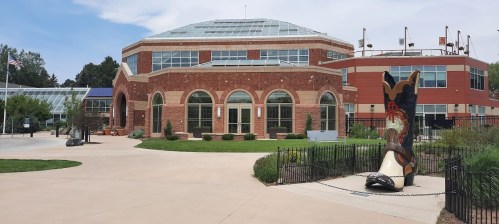 A modern brick building with large windows, surrounded by greenery and a giant cowboy boot sculpture in the foreground.