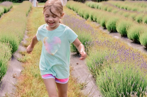 A young girl runs joyfully through a lavender field, surrounded by vibrant green and purple plants.