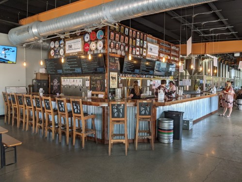 A spacious bar area with wooden stools, a metal counter, and a menu board, featuring patrons and staff inside a modern venue.
