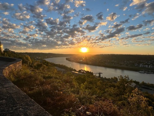 Sunset over a river, with clouds in the sky and greenery in the foreground, creating a serene landscape.