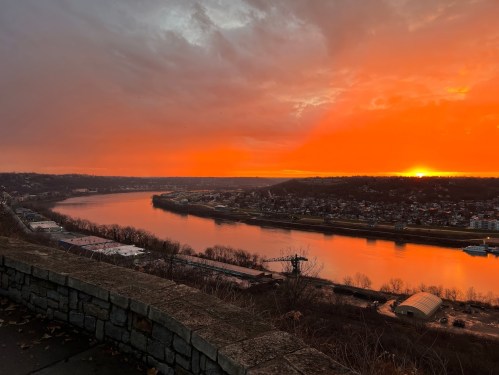 A vibrant sunset over a river, with clouds reflecting orange hues and a scenic view of the landscape and buildings.