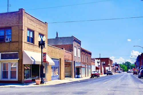A sunny street scene featuring brick buildings, storefronts, and a clear blue sky in a small town.