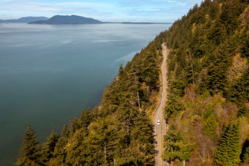 A winding road along a forested coastline with a calm sea and distant mountains under a clear blue sky.