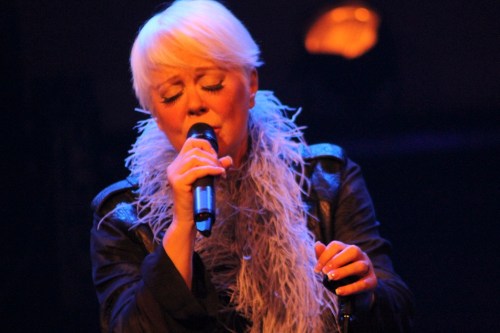 A singer with short blonde hair performs passionately on stage, wearing a black jacket and a feather boa.