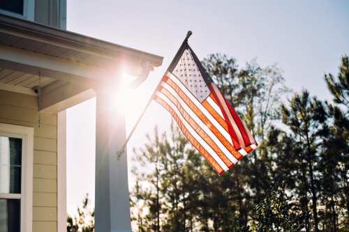 An American flag waves in the sunlight, with trees in the background and a clear blue sky.