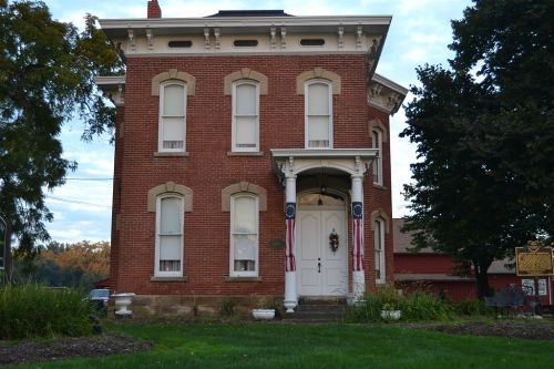 A historic brick house with white trim, featuring a porch and decorative elements, surrounded by greenery.