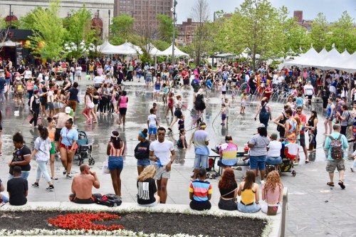 A vibrant outdoor scene with a large crowd enjoying a festival, featuring colorful tents and people dancing in a water area.
