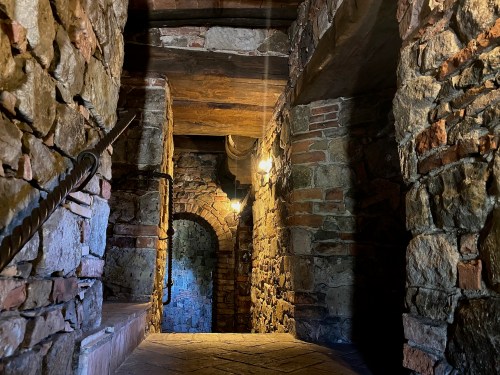 A dimly lit stone corridor with archways and wooden beams, featuring warm light from hanging lanterns.