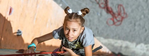 A young girl climbs a rock wall, focused and determined, with climbing gear and ropes in the background.