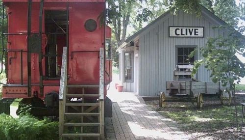 A red train car next to a small gray building with a sign reading "CLIVE," surrounded by greenery and a brick path.