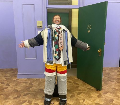 A person stands in a doorway, arms outstretched, wearing multiple layers of oversized clothing and a colorful tie.