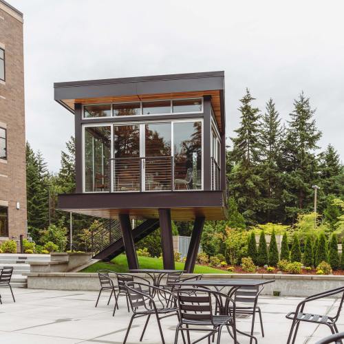 A modern glass-walled structure elevated on stilts, surrounded by greenery and outdoor seating.