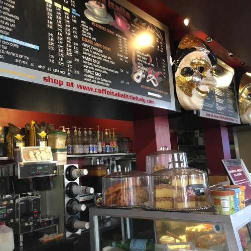 A café menu board with various food and drink options, featuring a decorative mask and display of cakes.