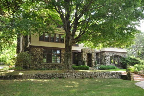 A large stone house surrounded by lush greenery and a prominent tree in the front yard.