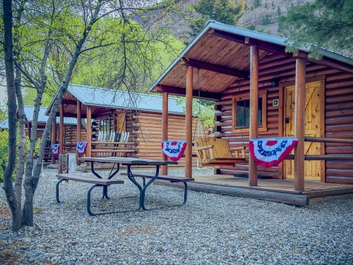 Two rustic log cabins with porches, surrounded by trees and gravel, decorated with red, white, and blue bunting.