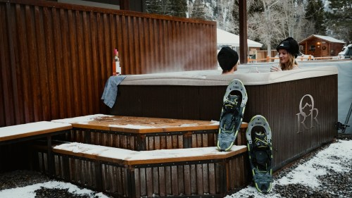 A couple relaxes in a hot tub surrounded by snow, with snowshoes nearby and a bottle of wine on the side.