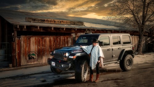 A person in a poncho stands next to a rugged black Jeep Wrangler in front of a rustic wooden building at sunset.