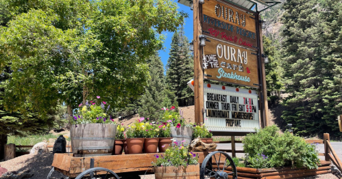 Sign for Ouray Steakhouse with flower pots and a rustic wagon, surrounded by trees and mountains.