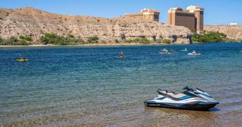 A jet ski sets along the shore of the Colorado River near Laughlin, Nevada. Kayakers and paddleboarders are visible on the water in the distance.