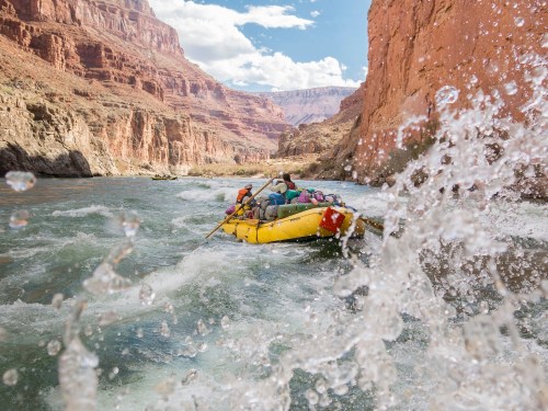 A group of people rafting through a rocky canyon, splashing water in a vibrant river under a blue sky.