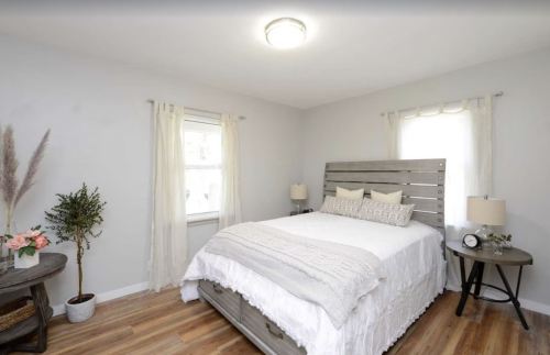 Cozy bedroom with a wooden bed, light gray walls, and natural light from a window, featuring plants and decorative pillows.