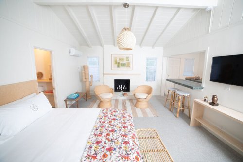 Bright, airy room with a bed, two chairs, a fireplace, and a small kitchen area, featuring light wood and floral decor.