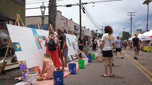 People painting on large canvases outdoors at a street festival, with tents and palm trees in the background.