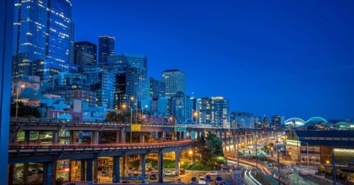 City skyline at dusk with illuminated buildings and busy roads, showcasing urban life and architecture.