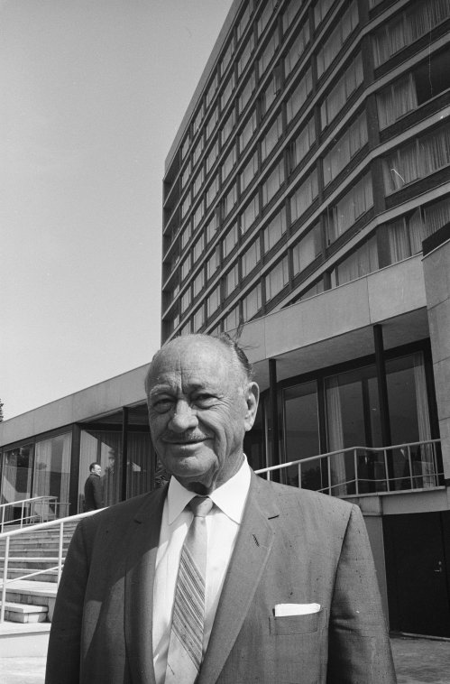 A man in a suit stands in front of a modern building, smiling at the camera on a sunny day.