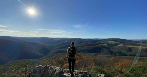 A person stands on a rocky outcrop, overlooking a vast landscape of rolling hills under a clear blue sky.