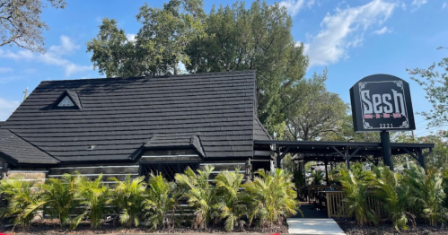 A rustic building with a black roof and a sign reading "Sesh," surrounded by palm trees and blue skies.