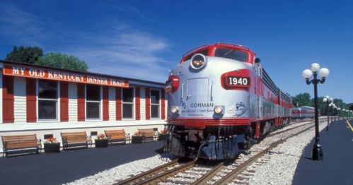 A vintage train at a station with a sign reading "My Old Kentucky Dinner Train" under a clear blue sky.